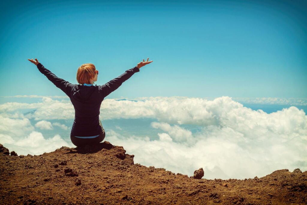 A woman uses practical applications and now enjoys a scenic view atop a cliff in Kula, Hawaii, surrounded by clouds and blue sky.