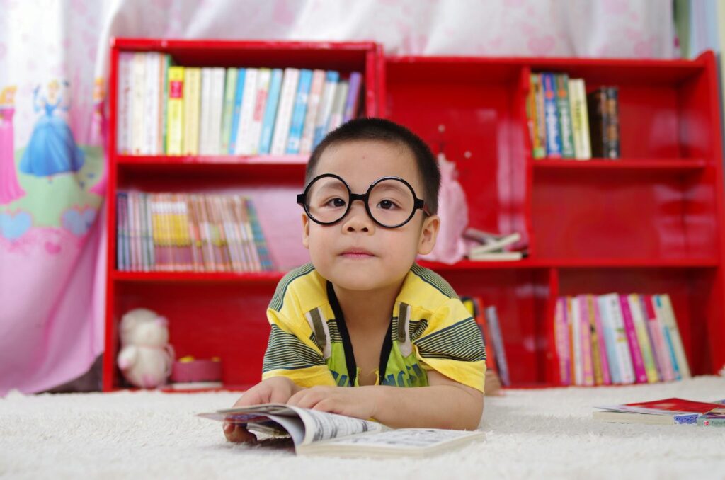 A cute child in glasses is reading a book about personal development surrounded by colorful shelves.