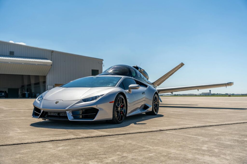 Silver Lamborghini parked beside a private jet at an airport in Illinois on a sunny day.