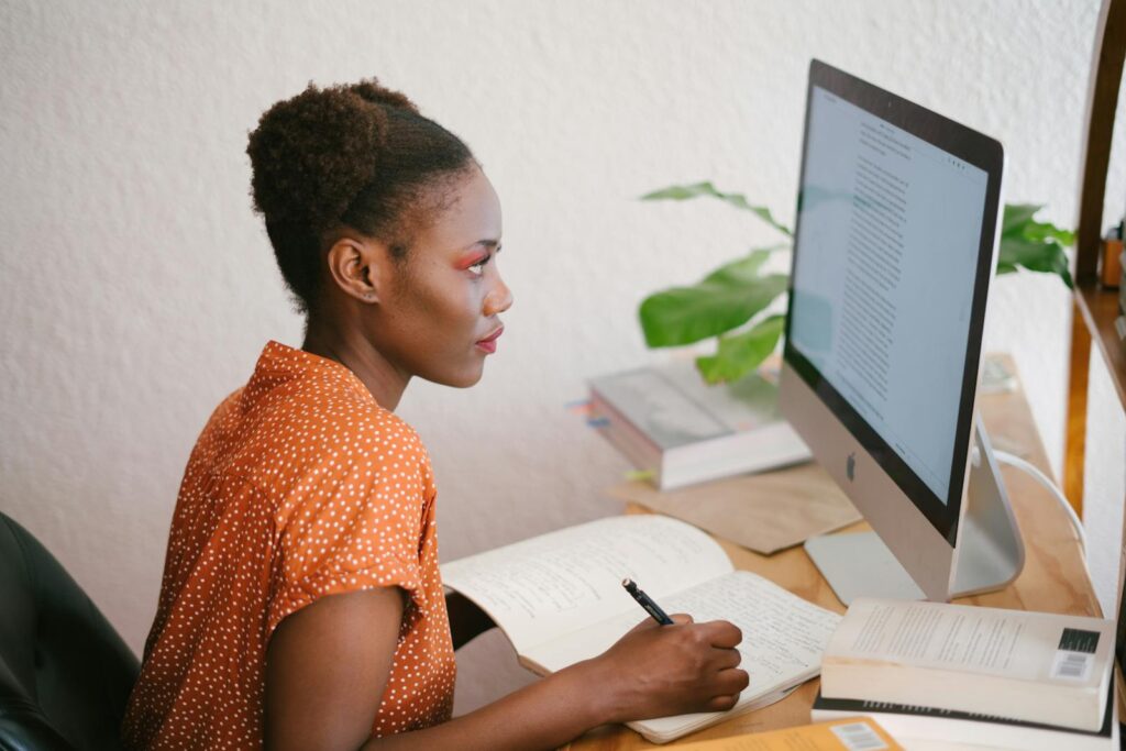 A focused young woman studying with books and an abundance mindset while on the computer at her home office desk.