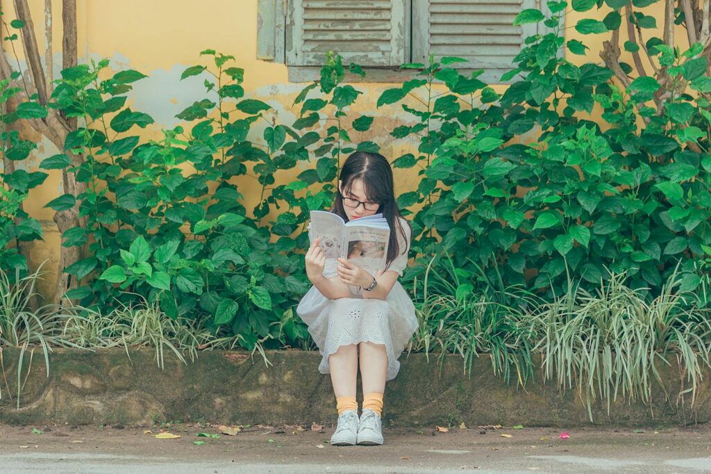A young girl sits on a bench outdoors, immersed in a book about personal development. Lush greenery surrounds her